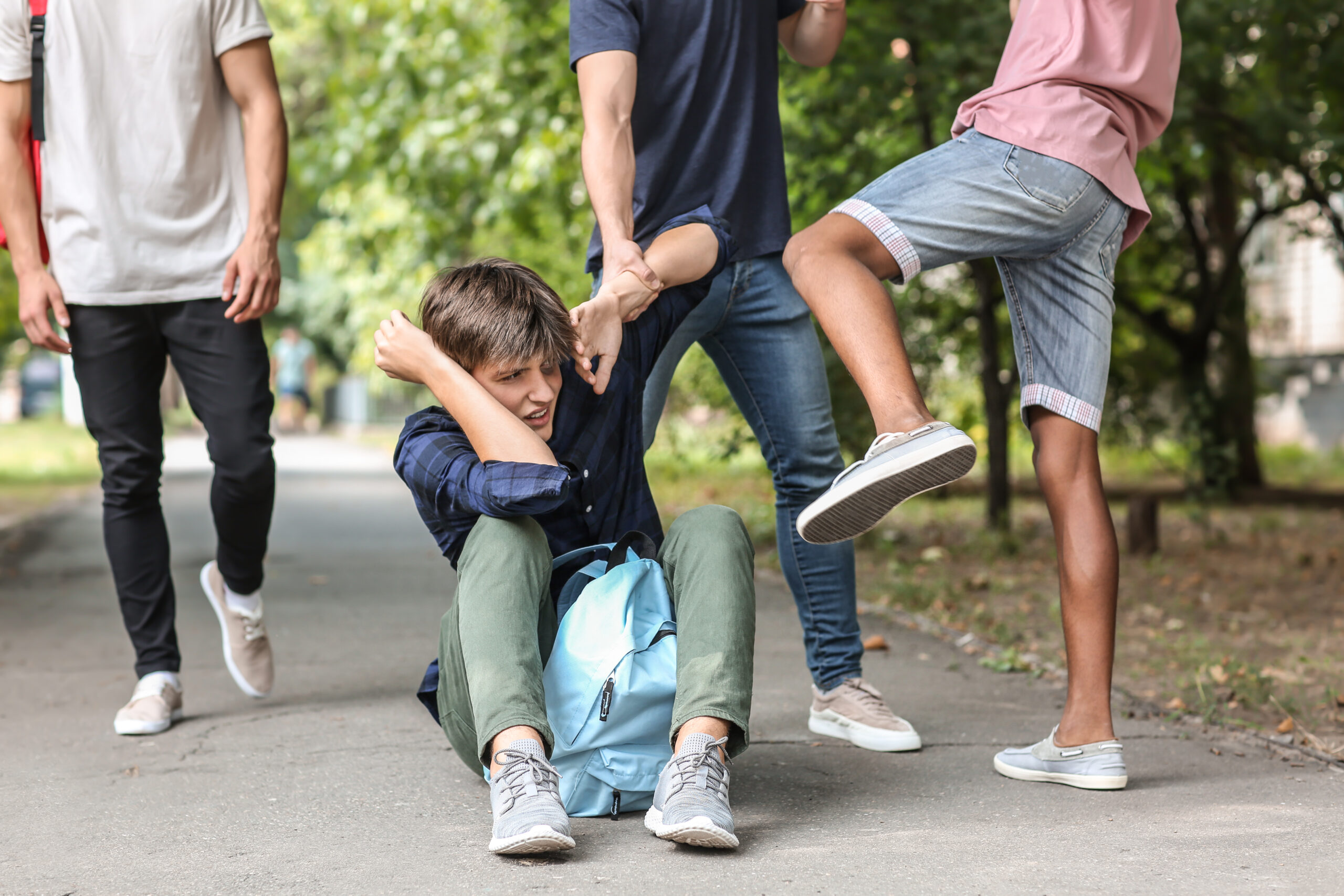 A group of teenagers bullying another teenager who is sitting on the ground with a backpack.