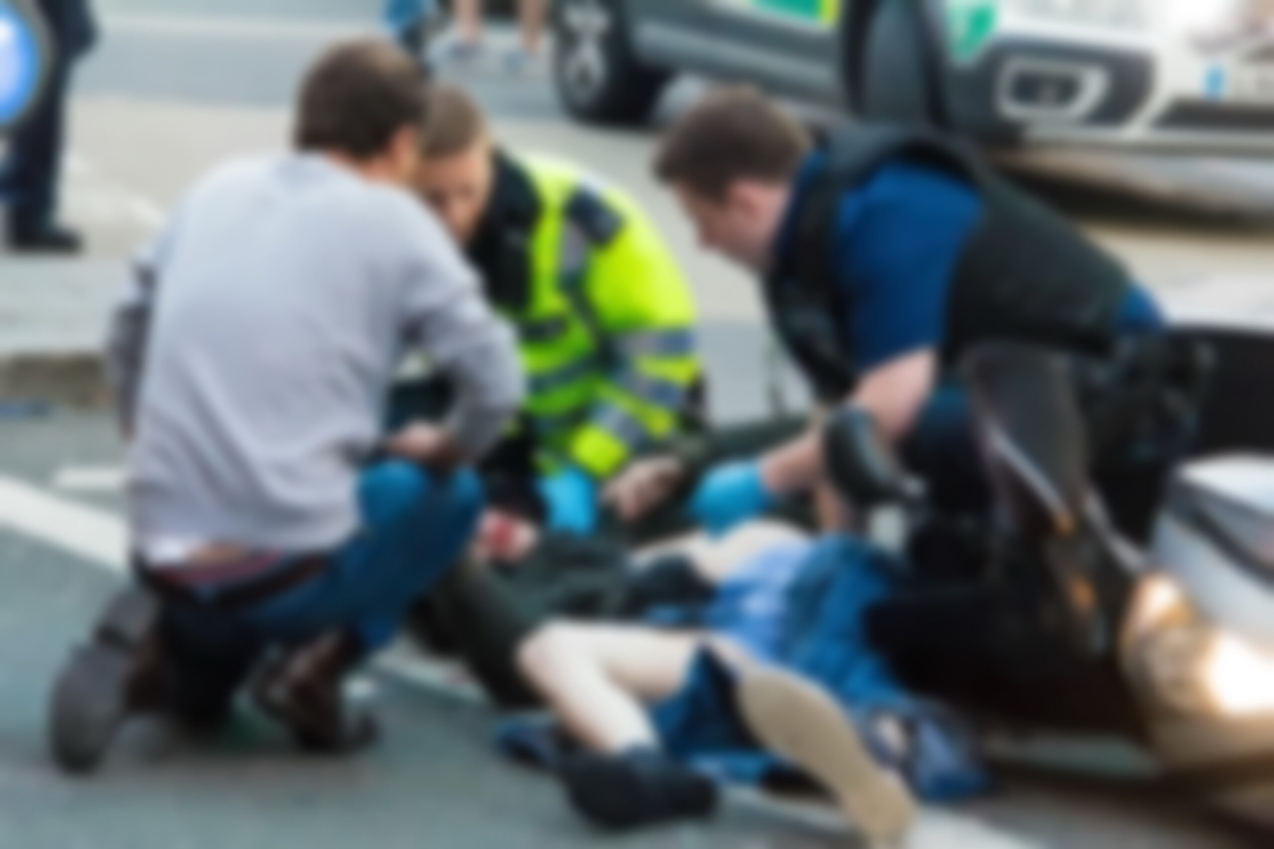 hree people, including two emergency responders, attending to an individual lying on the ground near a vehicle in an urban setting.
