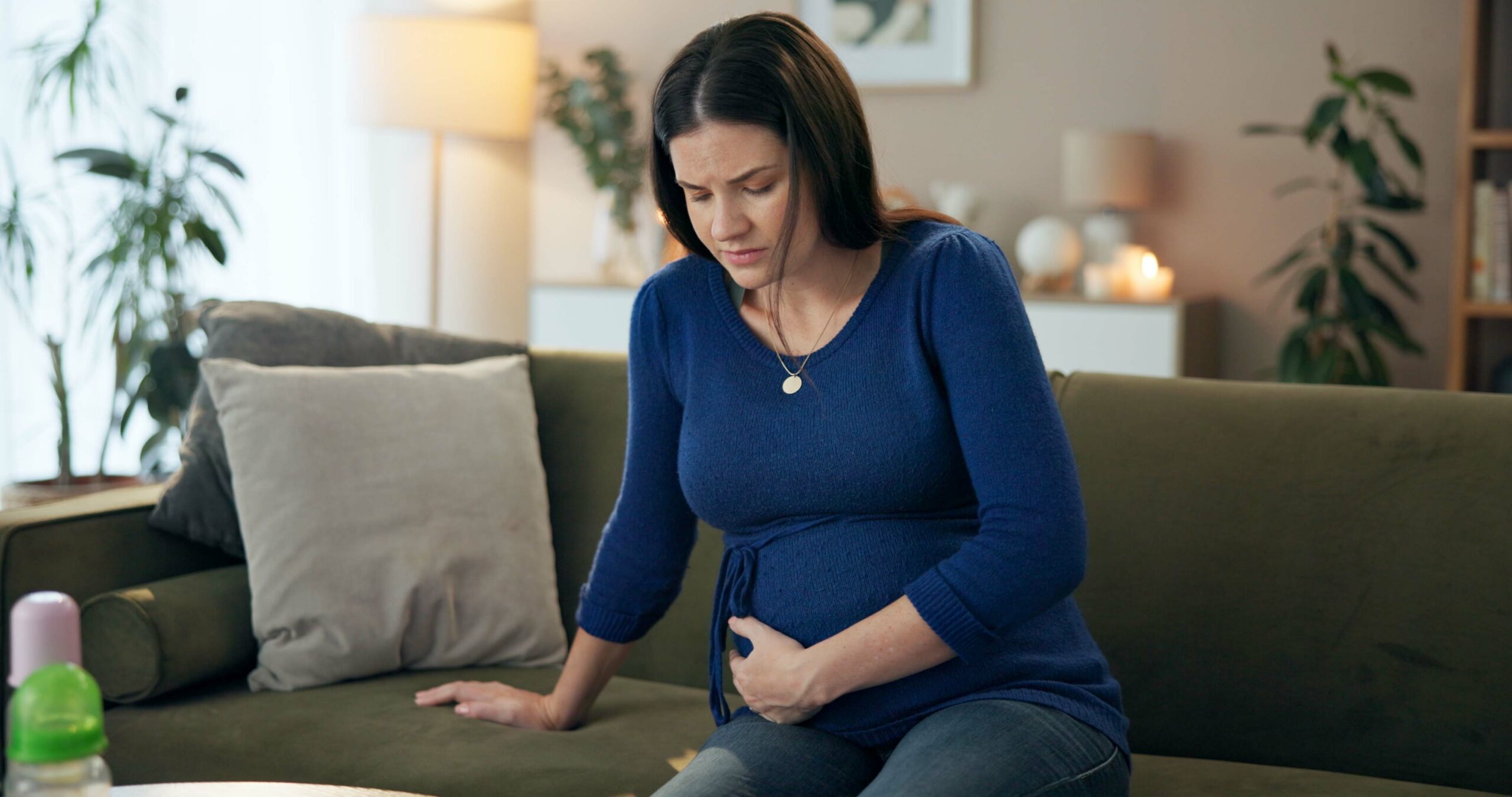 Pregnant woman experiencing abdominal pain during pregnancy while sitting on a couch at home.